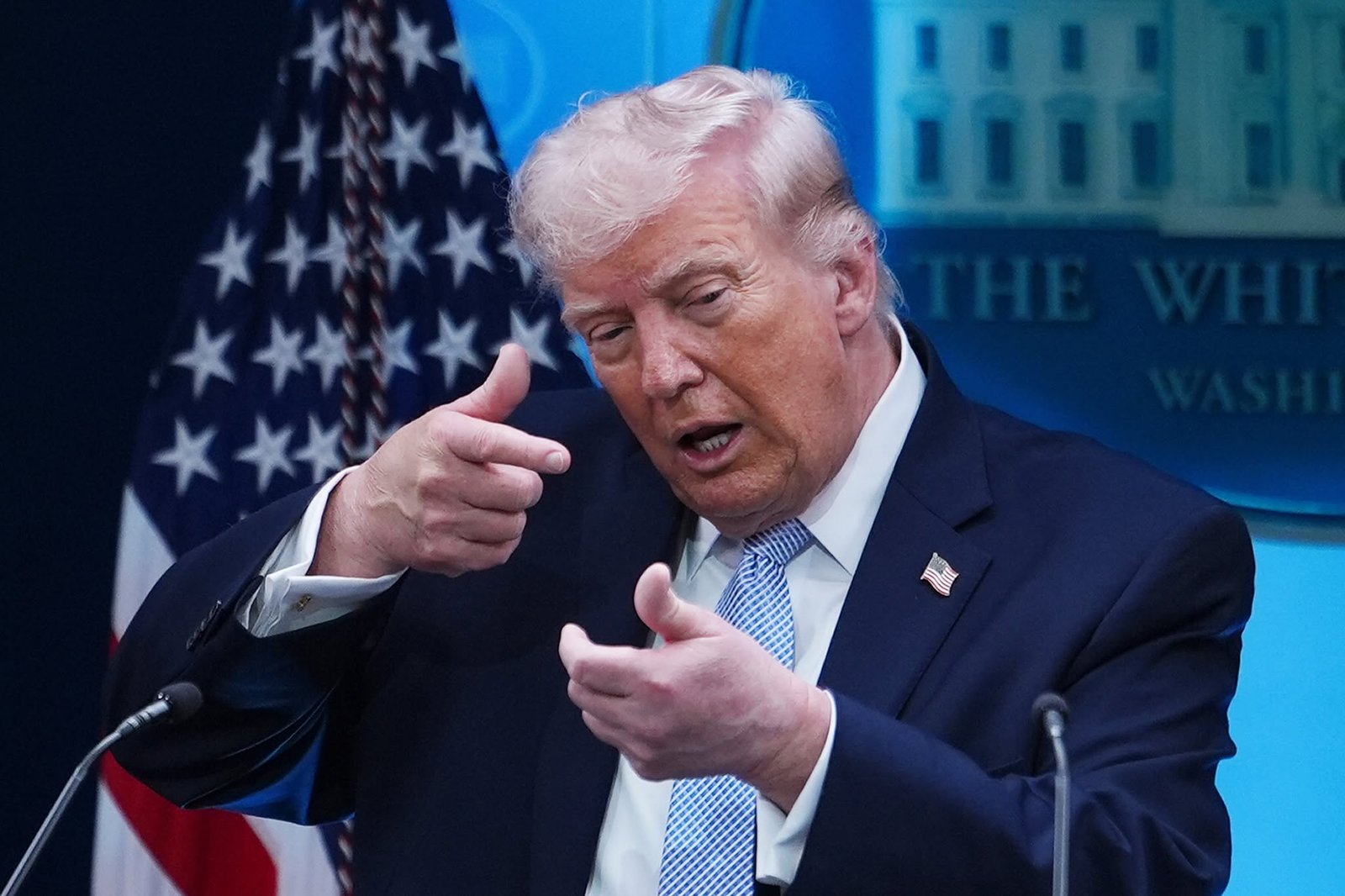 A man in a dark suit speaks at a podium in front of reporters in a briefing room.