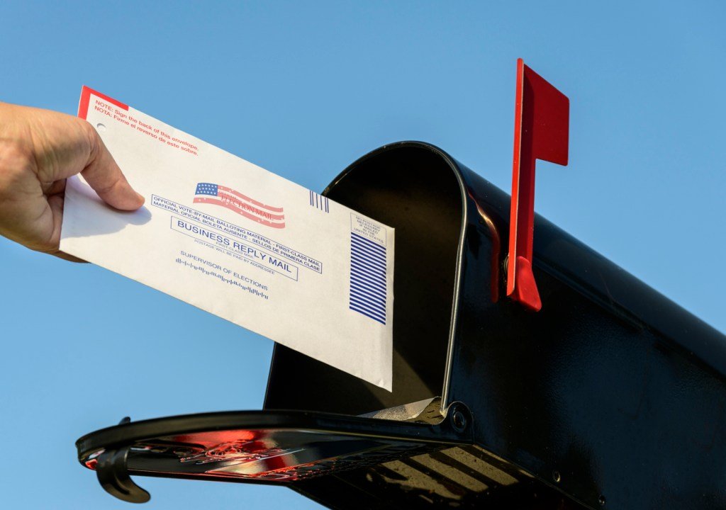 A person places a sealed election ballot envelope into a blue U.S. Postal Service collection box.
