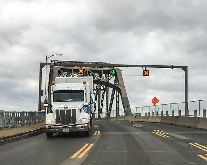 A commercial truck drives across a bridge connecting two countries, with border infrastructure visible.