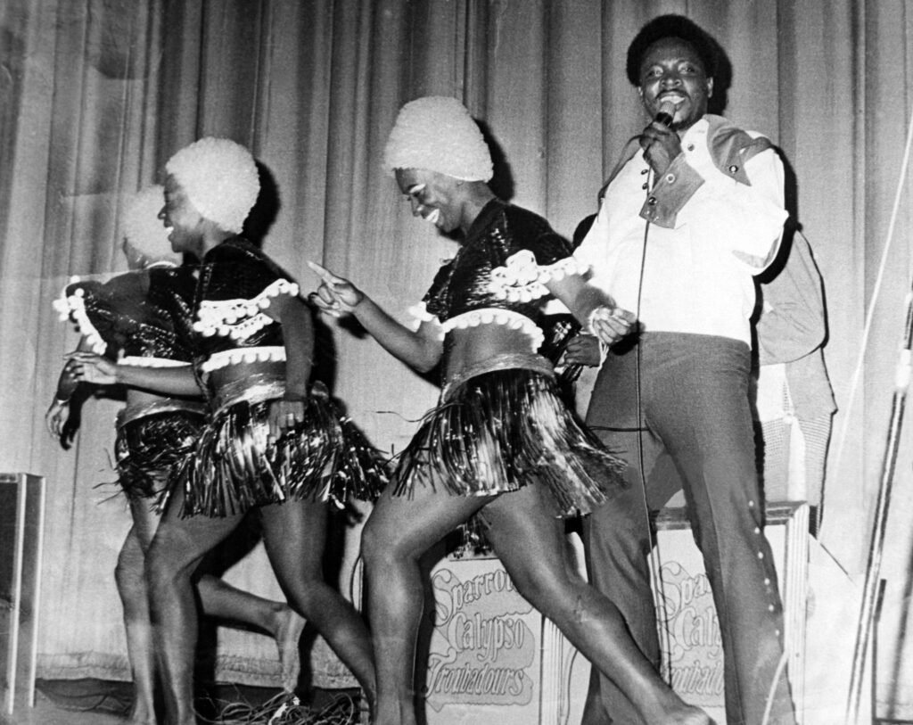 Archival stage photo of a calypso performer singing into a microphone during a live show.