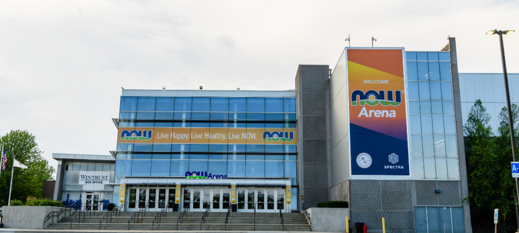The exterior of a large indoor arena with a parking lot in front and a cloudy sky above.