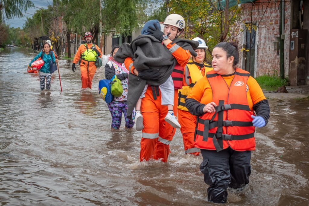 Residents wading through a flooded street lined with partially submerged houses.