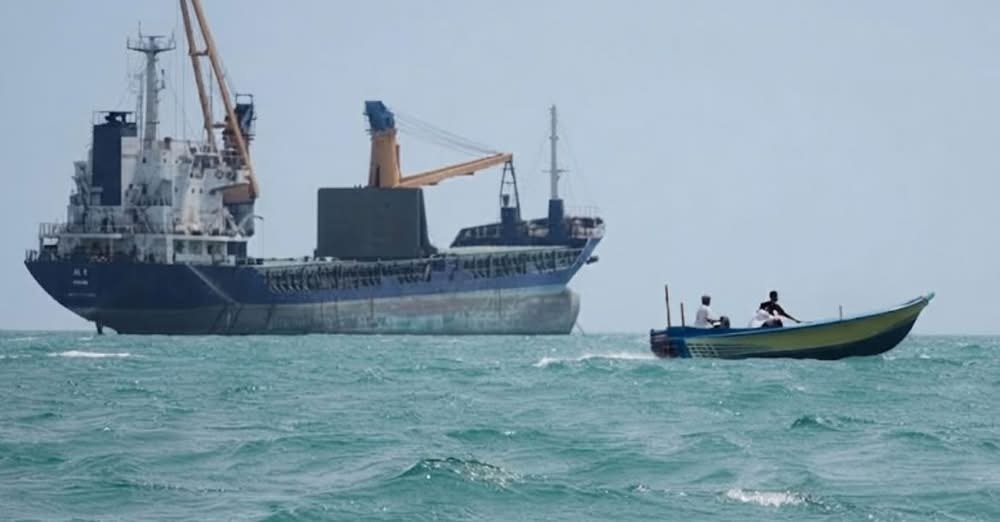A large cargo ship with cranes sits near a small boat carrying two people on open water.