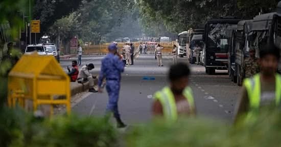 Uniformed police officers standing at a checkpoint on a Delhi street while commuters walk through.