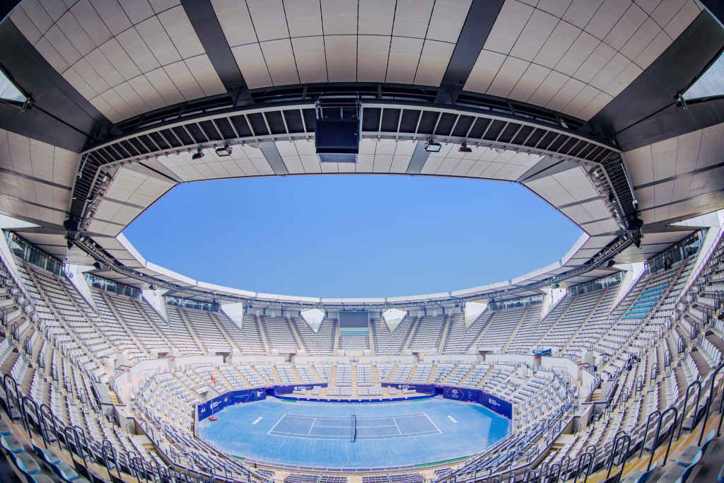 A large tennis stadium with an open roof and tiered seating surrounding a blue hard court.