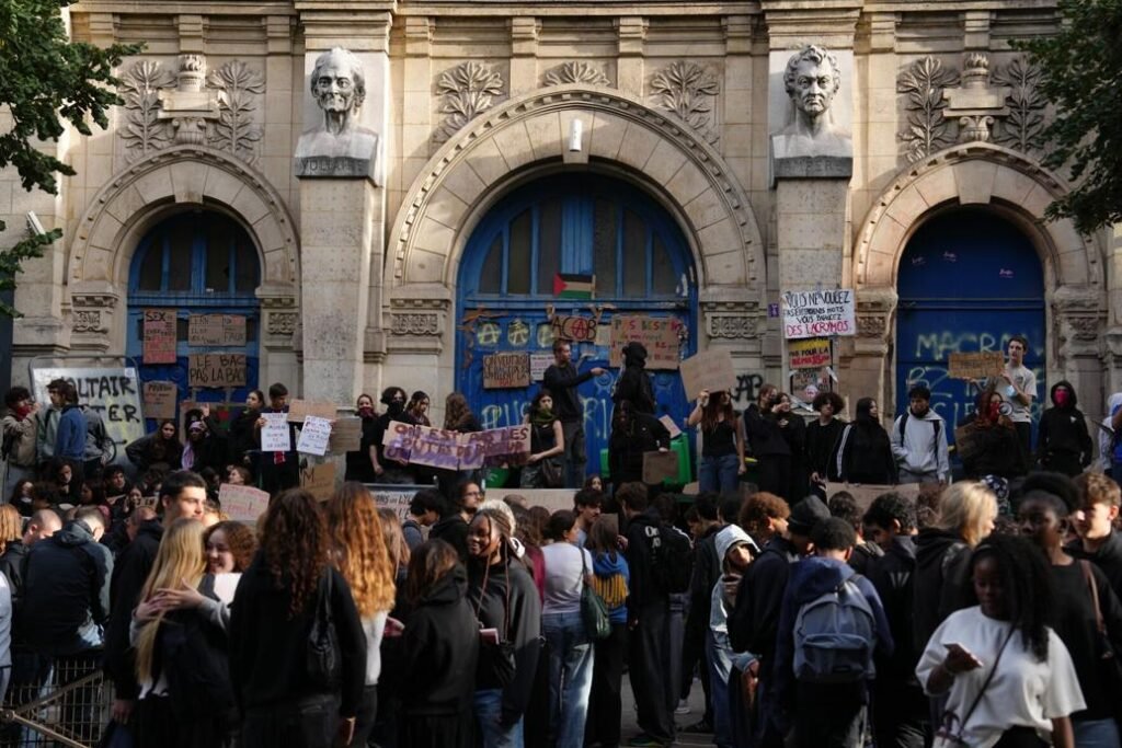 A crowd of teenage students gathers outside a school building during a demonstration.