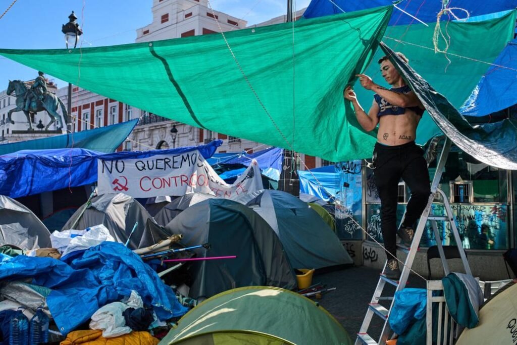 A man adjusting a tarpaulin over a tent at a protest camp in a Madrid square.