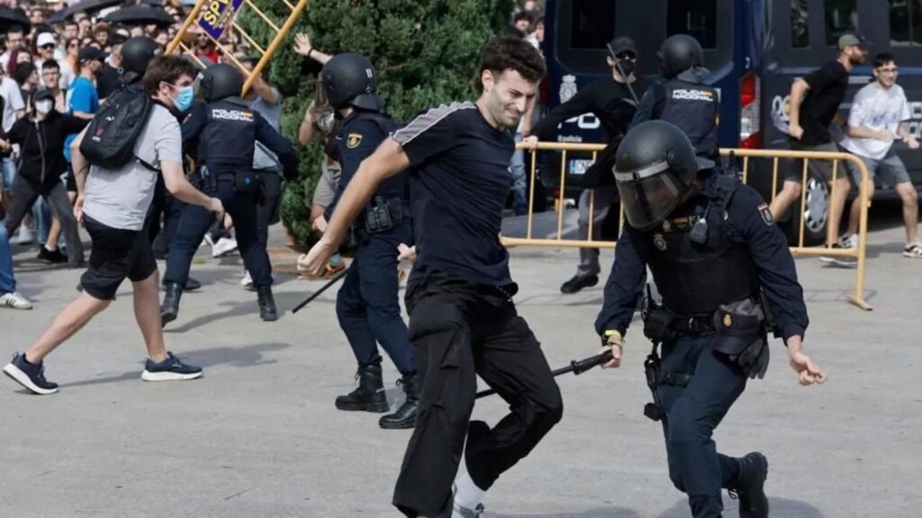 A protester runs as a police officer in riot gear reaches toward him, with other officers and a crowd visible nearby.
