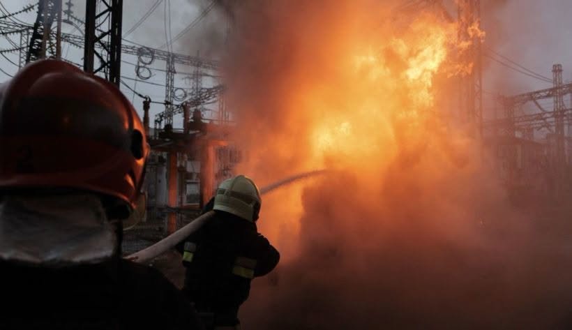 Firefighters and workers at a damaged power facility with smoke and debris.