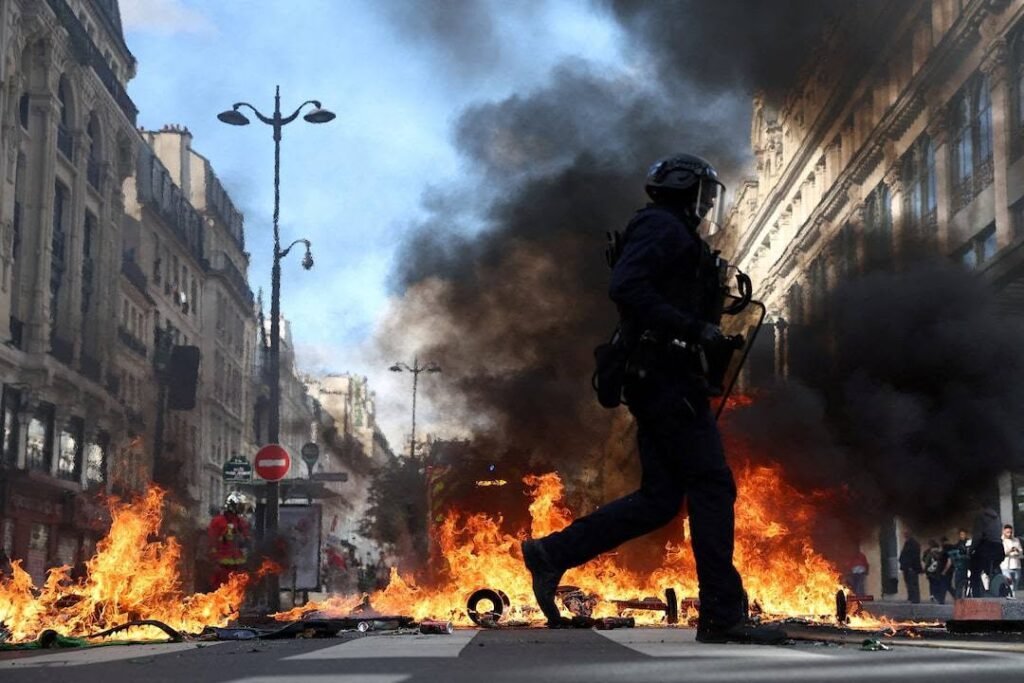 Riot police in helmets and body armor walking past a street barricade on fire.