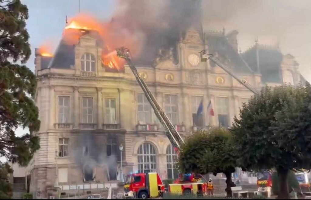 Firefighters spraying water at a historic building with flames and smoke rising from its roof.