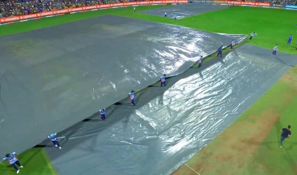 Ground staff covering a cricket pitch under rain with floodlights on at a stadium.