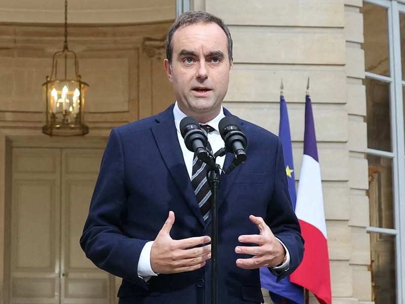 A man in a dark suit speaking at a lectern in front of French flags.