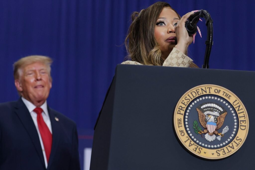 A woman in a gold suit speaking at a podium at a political rally.