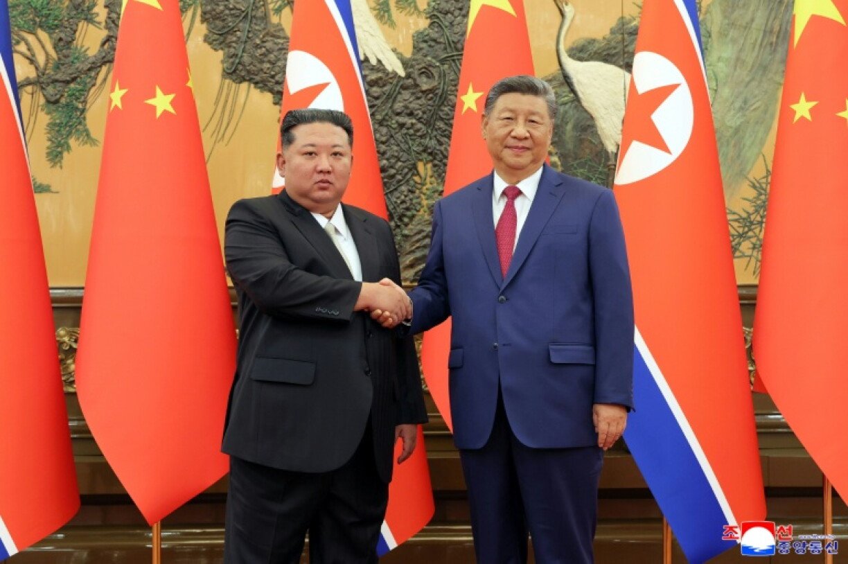 Two men in dark suits shaking hands in front of flags.