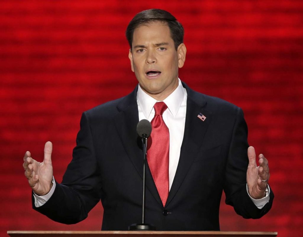 Marco Rubio speaking at a podium in a dark suit.