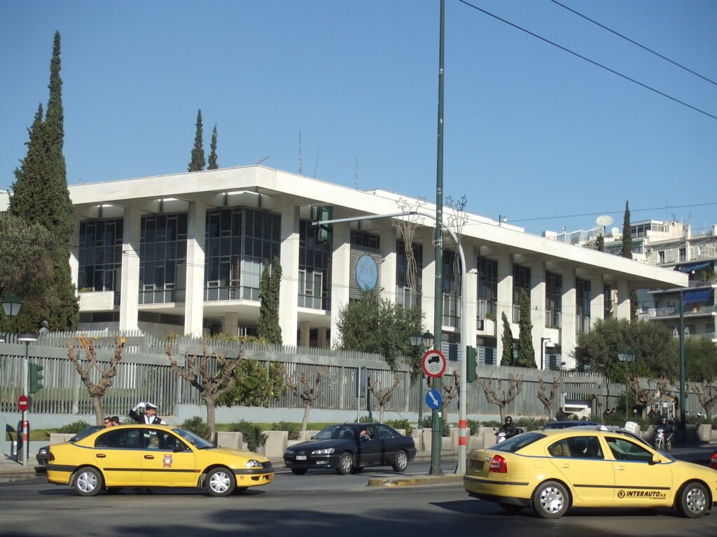 Front of a government office building flying the American flag, with a security perimeter and trees in the foreground.