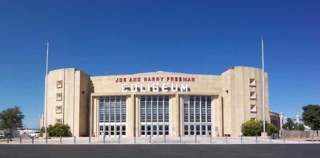 A large concrete arena building with a curved facade and a parking lot in front of it under a clear sky.
