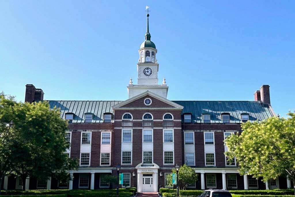 A brick academic building with a clock tower, surrounded by lawns and trees on a clear day.