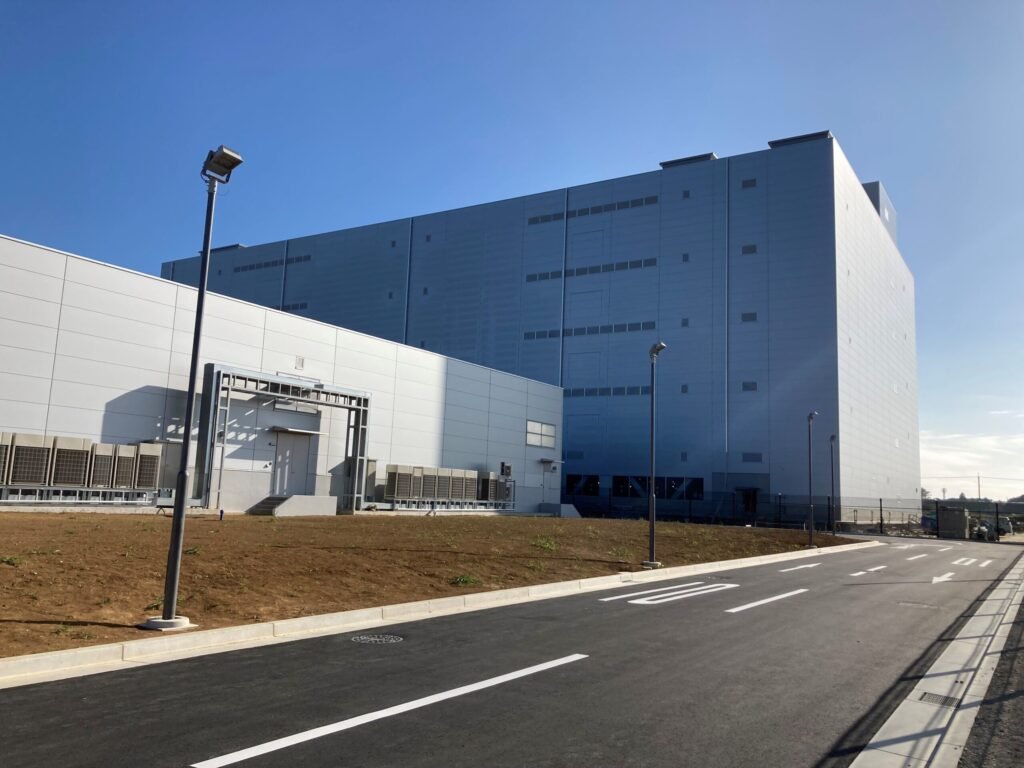 A large grey-blue windowless building with metal panel walls stands beside a lower white building with rows of outdoor cooling units, next to an empty road under a clear blue sky.