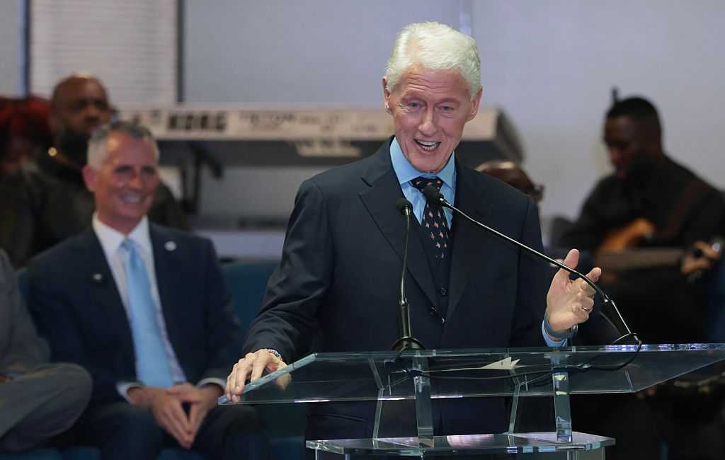 Bill Clinton speaking to a crowd inside a church hall while David Jolly sits behind him.