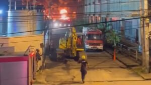Firefighters and fire trucks near a yellow excavator at night, with flames and a large plume of dark smoke rising behind buildings.