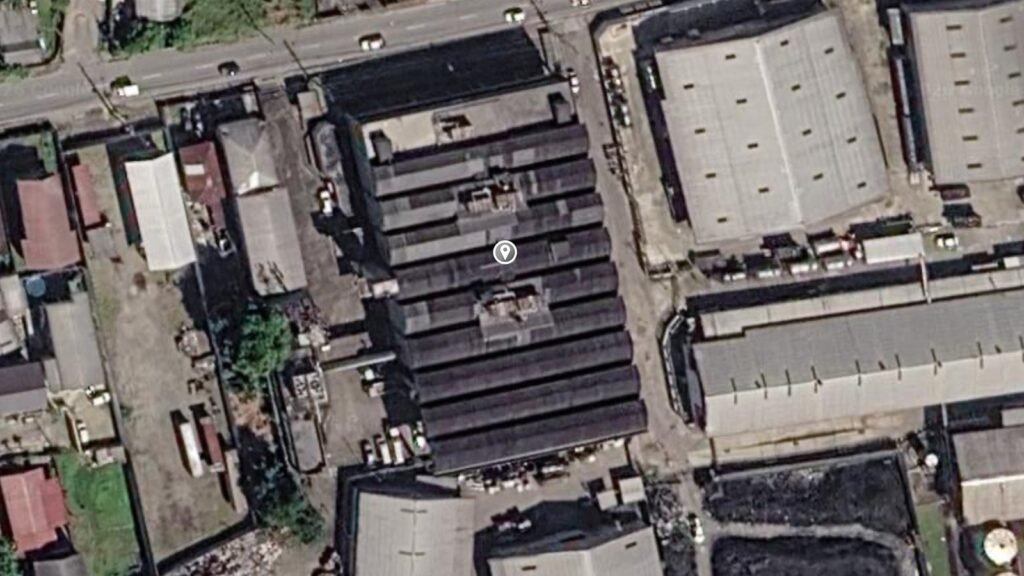 A satellite image of a large dark sawtooth-roofed building with a map pin, surrounded by warehouses, a road across the top and houses to the north and west.