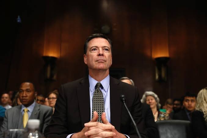 James Comey in a dark suit seated at a hearing table with microphones.