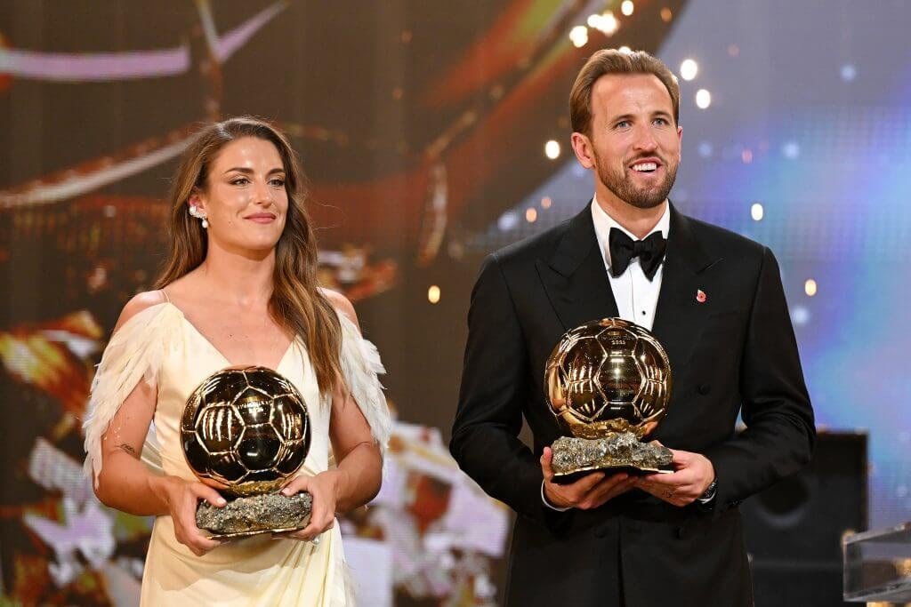 Two people stand on a stage, each holding a golden Ballon d’Or trophy: a woman in a cream gown with feathered shoulders and a man in a black tuxedo with a bow tie.
