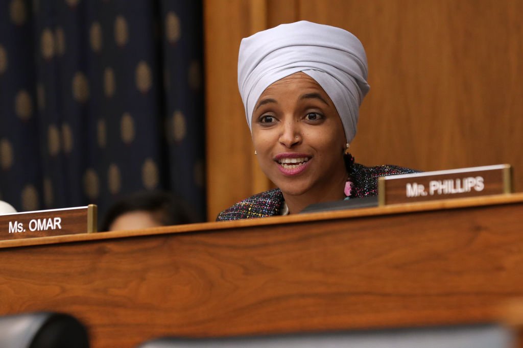 Ilhan Omar, wearing a head covering and a dark jacket, speaking at a microphone during a forum on Capitol Hill.