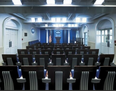 Rows of blue chairs face a podium in a wood-paneled briefing room.