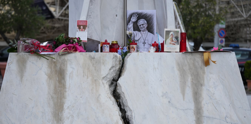 Candles and flowers placed around a framed portrait at the base of a statue outside a hospital.