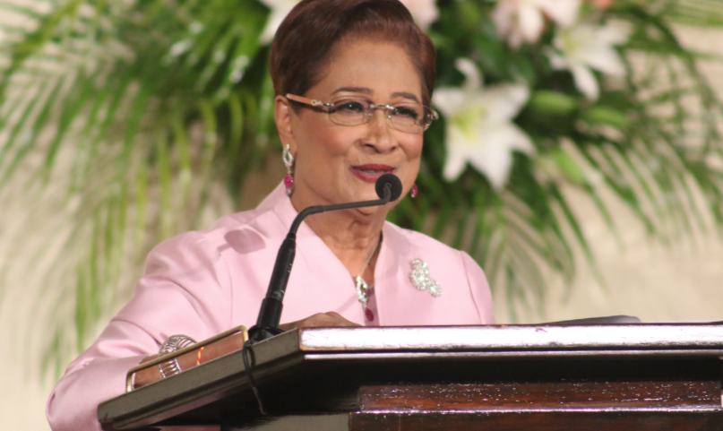 Kamla Persad-Bissessar in a bright outfit speaking at a podium.
