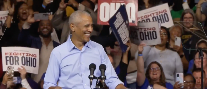 Barack Obama in a white shirt speaking at a podium in front of a large crowd.