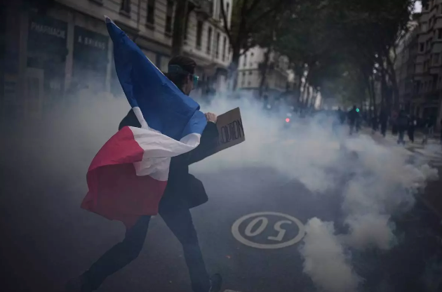 A person holding a flag walking through a cloud of tear gas on a street.