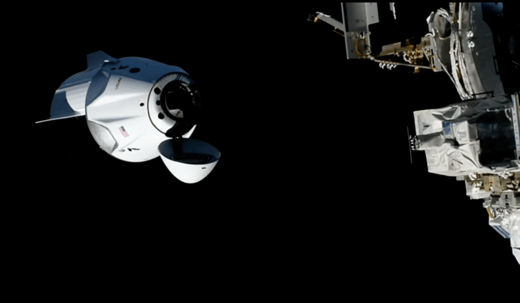 A white spacecraft with its nose cone open nears the space station above Earth.