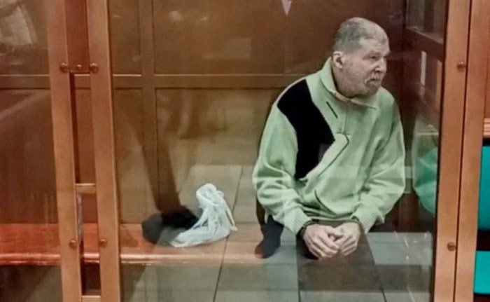 An older man standing in a glass courtroom enclosure.