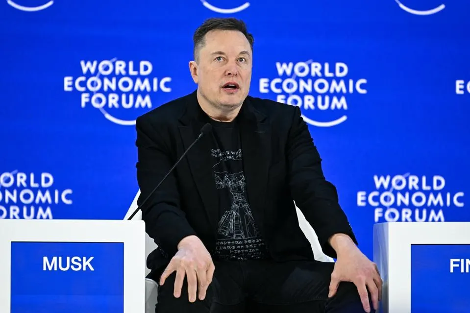 Elon Musk in a dark jacket speaking on a stage.