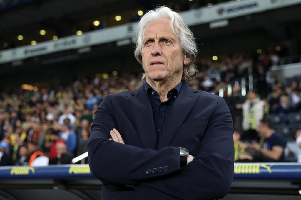A man in a dark jacket standing on the sideline of a football pitch.