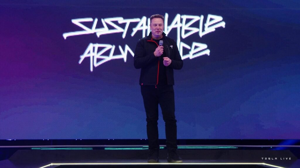 A man in a dark jacket speaking on stage in front of a screen.