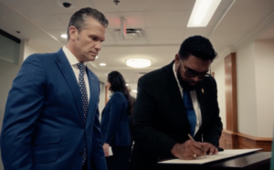Two men in dark suits signing documents at a table with flags behind them.