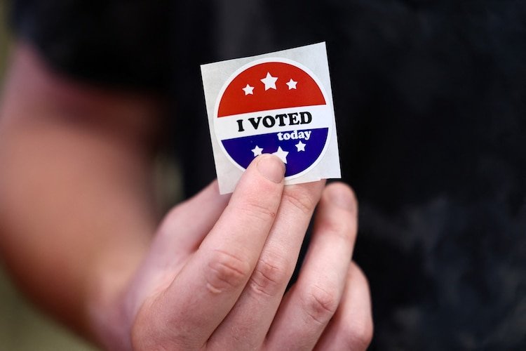 A hand holding an “I Voted” sticker inside a town hall during early voting.