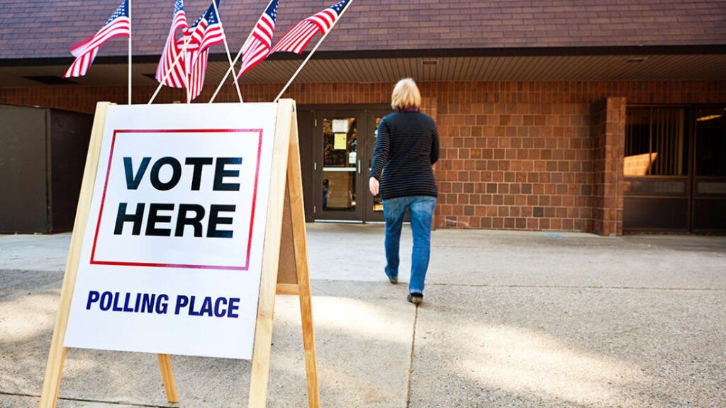 A “Vote Here” sign outside a building with American flags.