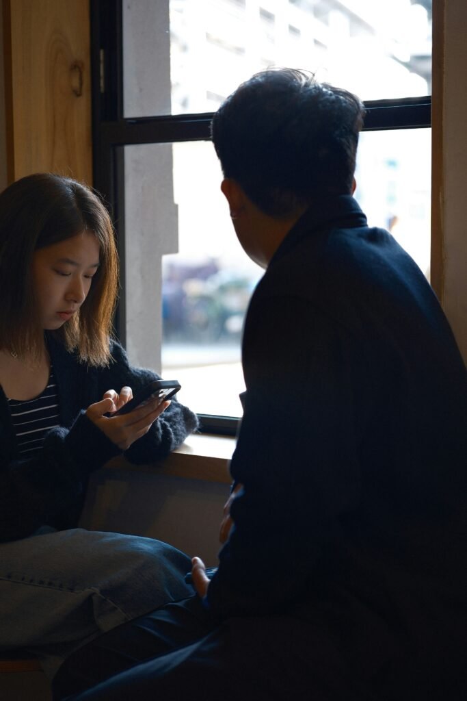 Young adult checking a smartphone while sitting beside a friend during an in-person social interaction.